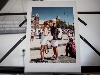 Photographic print of a couple kissing each other in front of the Amsterdam Rijksmuseum. The man of the couple is taking a selfie with his mobile phone while kissing. © Renske Vrolijk