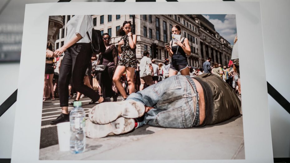 Print van voorbijgangers die kijken naar een dakloze man die kennelijk onverstoorbaar op de stoep ligt te slapen in Regent Street in Londen.