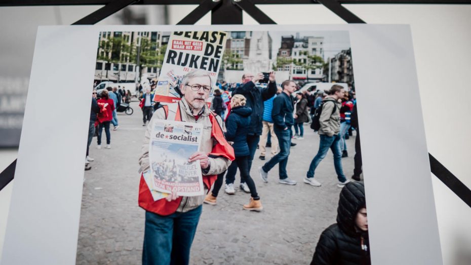 Print van een demonstrant op De Dam in Amsterdam. Hij verkoopt de krant De Socialist. Achter hem is een protestbord zichtbaar: "belast de rijken".