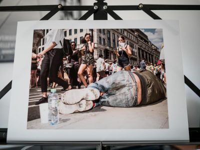 Print of passers-by looking at a homeless man who is apparently undisturbed asleep on the pavement on Regent Street in London.