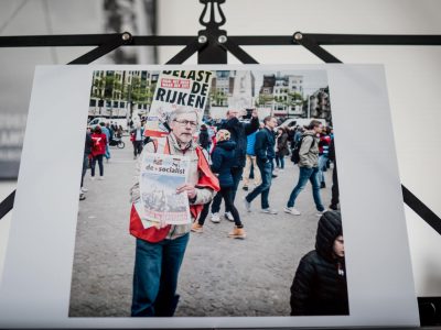 Print of a demonstrator on Dam Square in Amsterdam. He sells the newspaper De Socialist. Behind him a protest sign is visible: "Tax the rich".